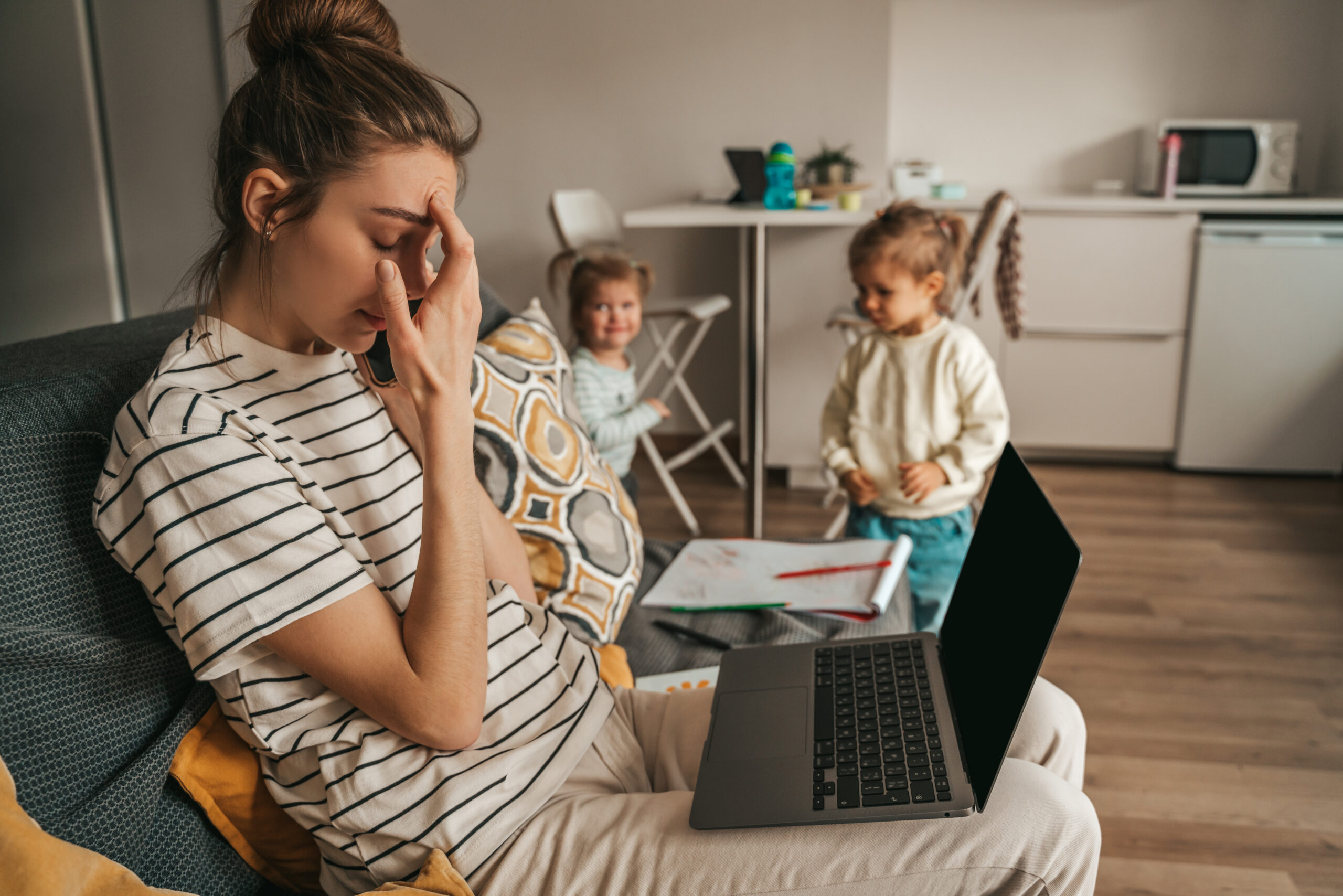 Tired housewife with the laptop and smartphone sitting on the sofa while her daughters playing